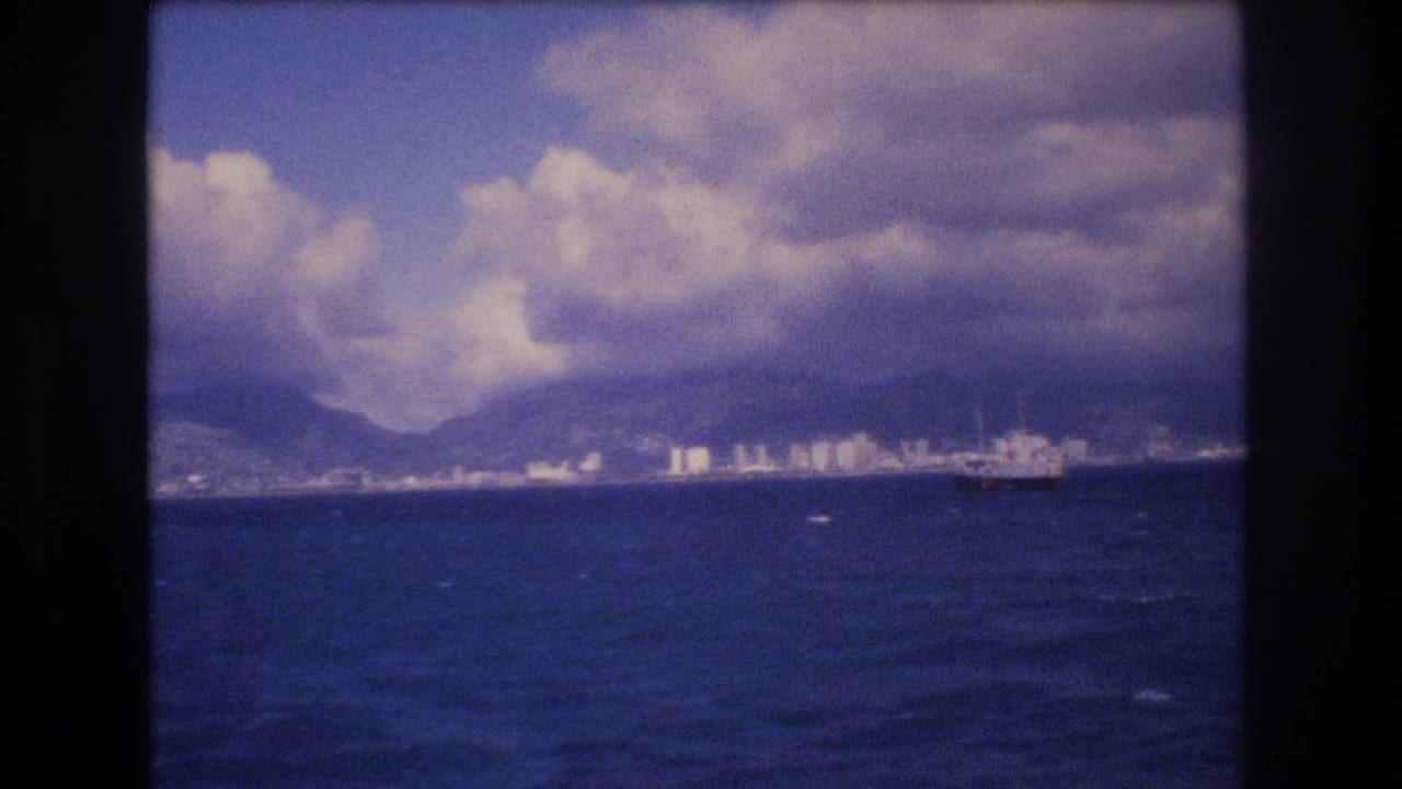 1977 Honolulu shoreline seen from across the ocean, with a distant ship, white buildings, mountains, and heavy clouds in a soft 8mm frame.