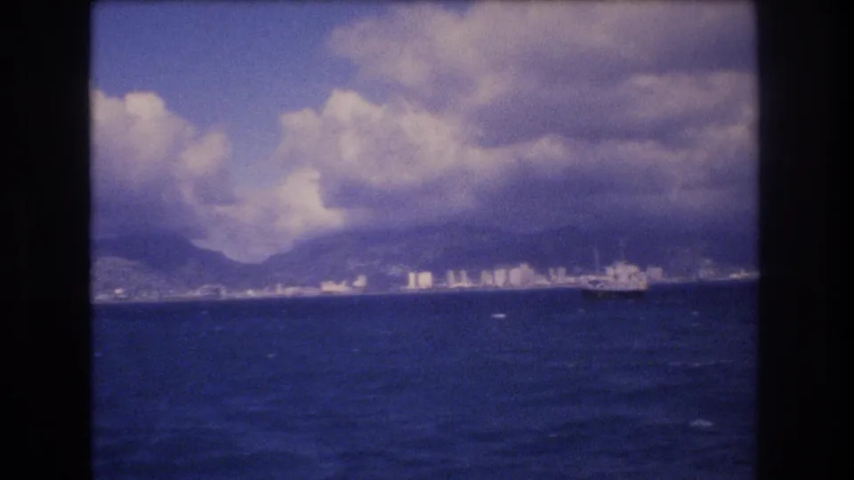1977 Honolulu shoreline seen from across the ocean, with a distant ship, white buildings, mountains, and heavy clouds in a soft 8mm frame.