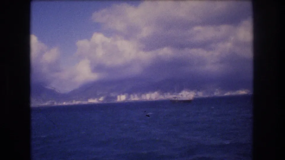 Distant Honolulu coastline across dark blue water, with white shoreline buildings, cloud-covered mountains, and a small vessel shape offshore.