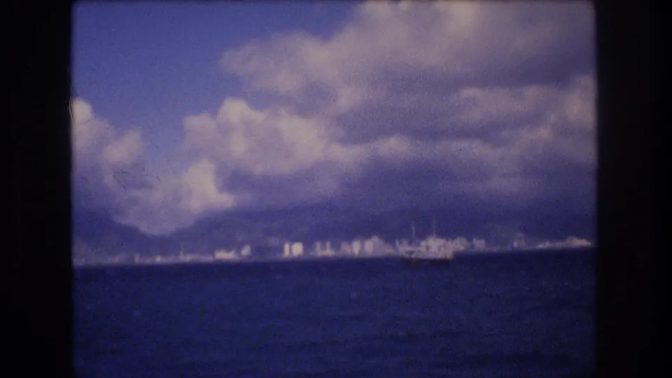 Soft, grainy 8mm frame of Honolulu from the water, showing blue ocean foreground, low city buildings, mountains, and a broad cloud bank.