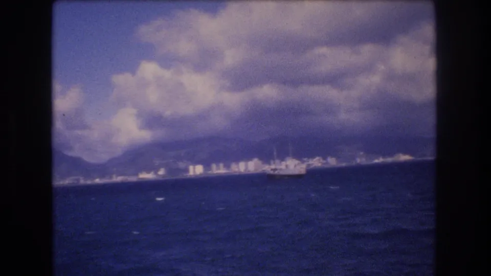 1977 Honolulu coastal view with a distant naval-looking ship on the water, shoreline buildings below mountains, and bright clouds across the sky.