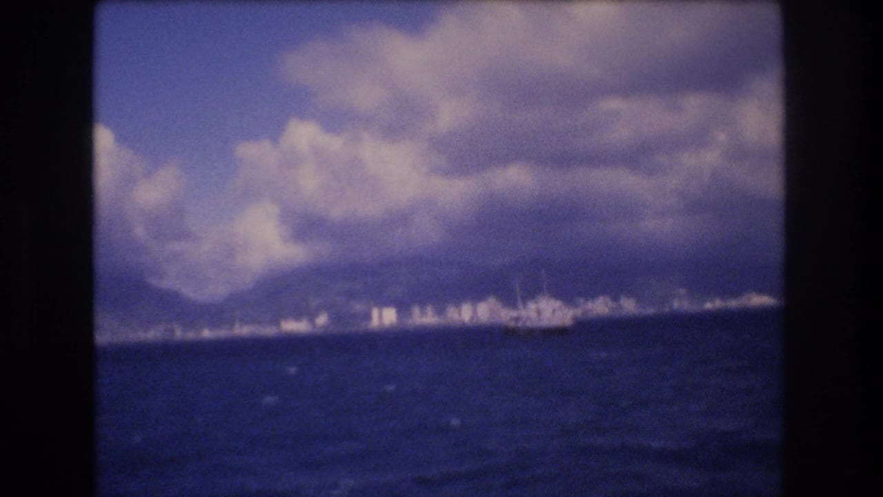 1977 8mm view from offshore toward Honolulu, with a large boat near the distant city shoreline, mountains, and heavy cloud cover.