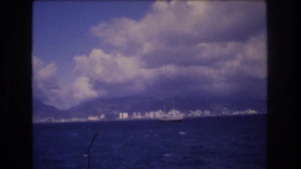 Distant 1977 Honolulu coastal view with ocean in the foreground, a large boat near shore, city buildings, mountains, and layered clouds.