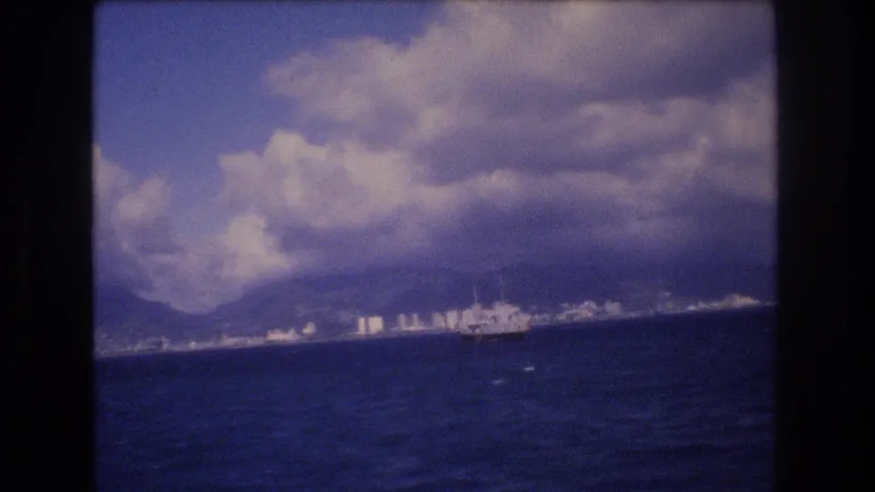 Soft 8mm frame of Honolulu from offshore in 1977, showing waves, a distant vessel, waterfront buildings, mountains, and bright cloud formations.