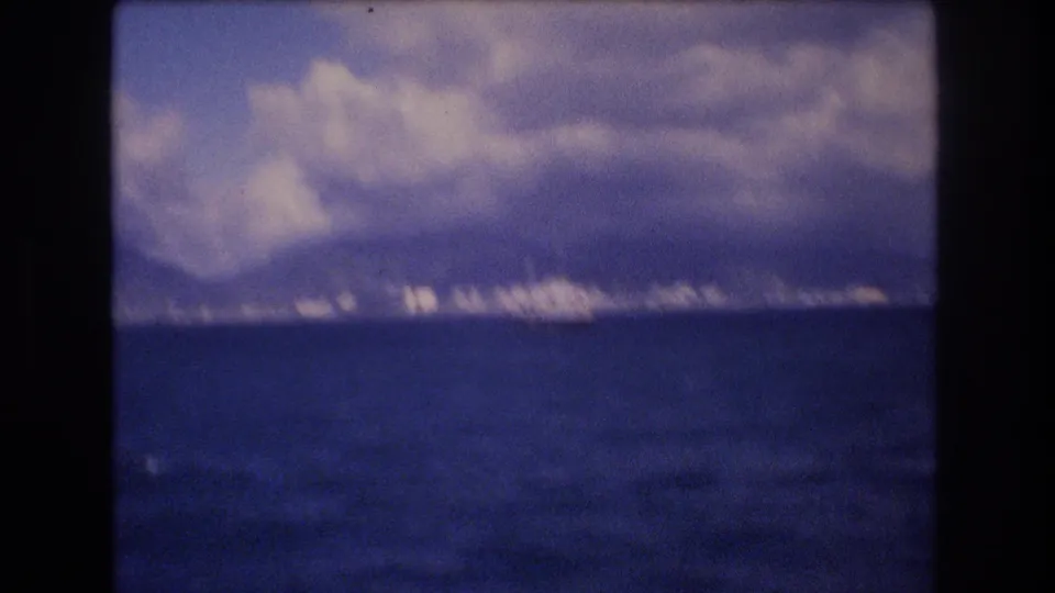 Soft, slightly blurred 1977 8mm view toward the Honolulu waterfront, with open ocean, a distant boat, city buildings, mountains, and heavy clouds.