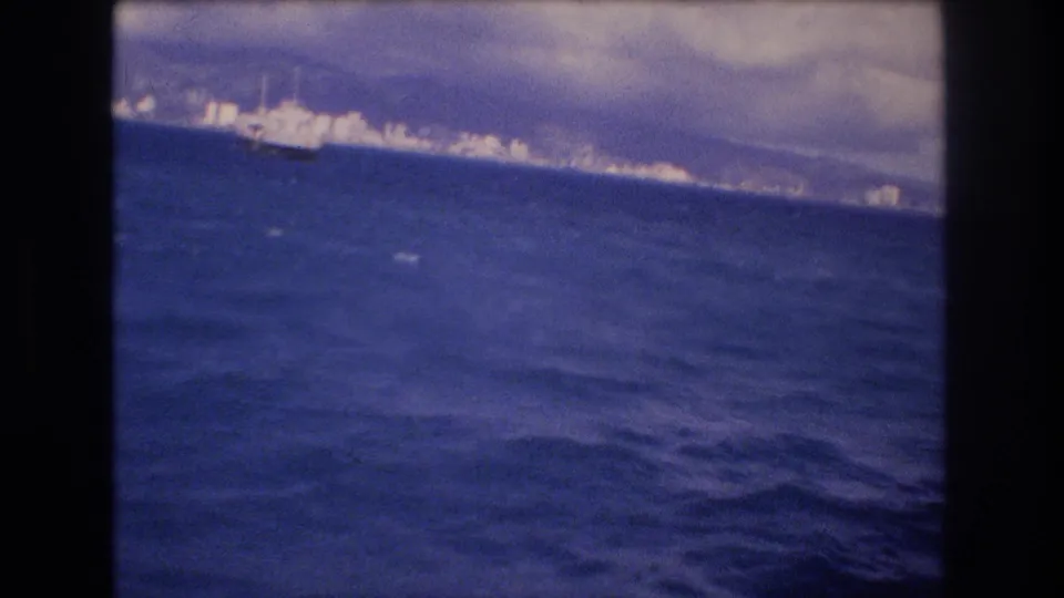 Distant Honolulu shoreline appears across the water in a 1977 8mm frame, with a large ship-like vessel and cloudy sky visible near the horizon.