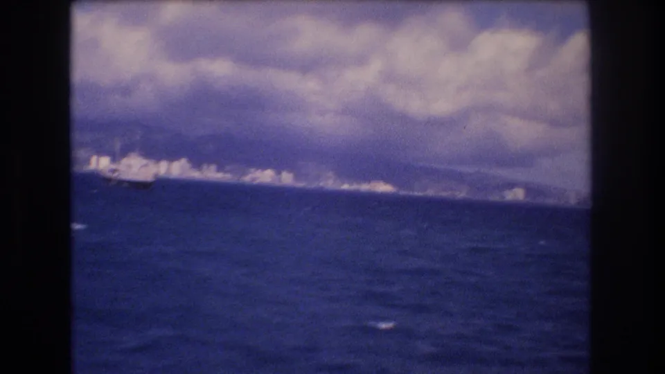 1977 Honolulu, Hawaii appears in the distance across blue ocean water, with a cloudy purple-tinged sky and dark 8mm frame borders.