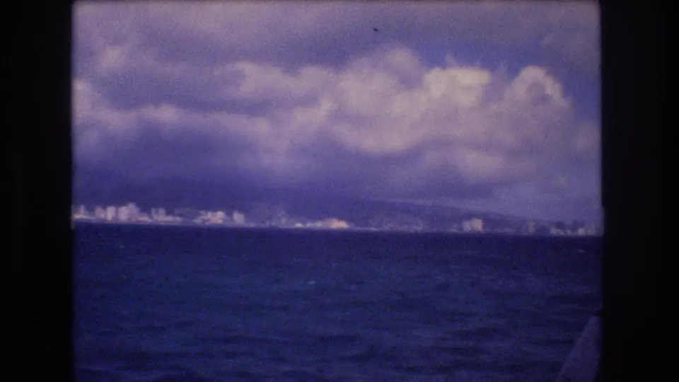 The frame holds on a distant Honolulu coastal strip, with low white buildings, possible hills behind them, and heavy clouds above the ocean.