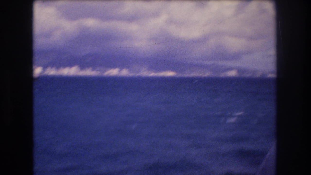 1977 Honolulu-area 8mm frame showing blue ocean from a moving boat, with a distant low shoreline and heavy clouds above the horizon.