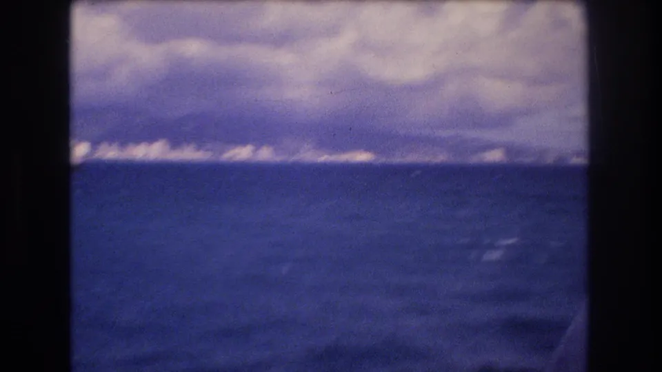 1977 Honolulu-area 8mm frame showing blue ocean from a moving boat, with a distant low shoreline and heavy clouds above the horizon.