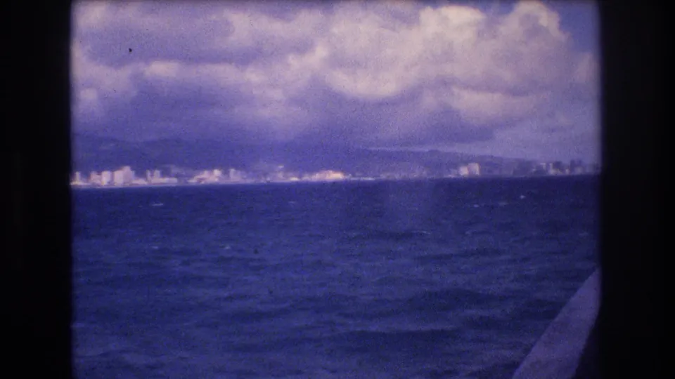1977 Honolulu-area coastal view from offshore, with white shoreline buildings, mountain ridges, choppy water, clouds, and part of the boat at right.