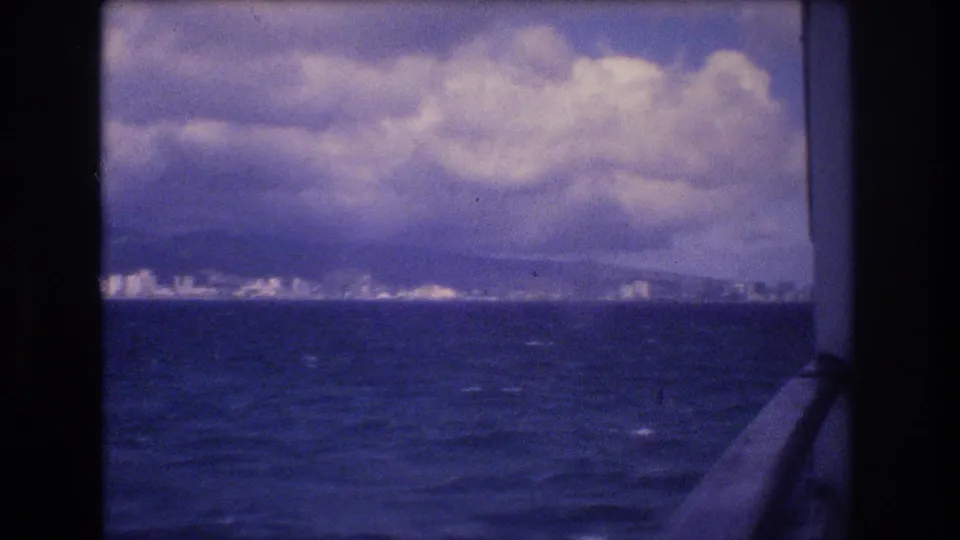 1977 Honolulu-area 8mm frame with a distant band of shoreline buildings below mountains and large clouds, viewed from a boat over choppy water.