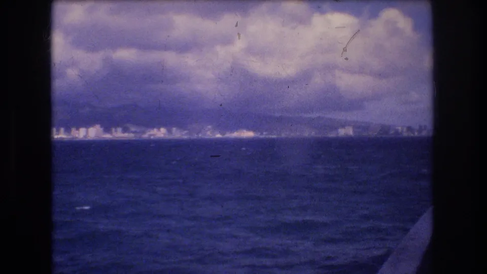 1977 Honolulu-area boat-trip frame showing choppy ocean, a distant city shoreline, mountain backdrop, heavy clouds, and the vessel edge along the right side.