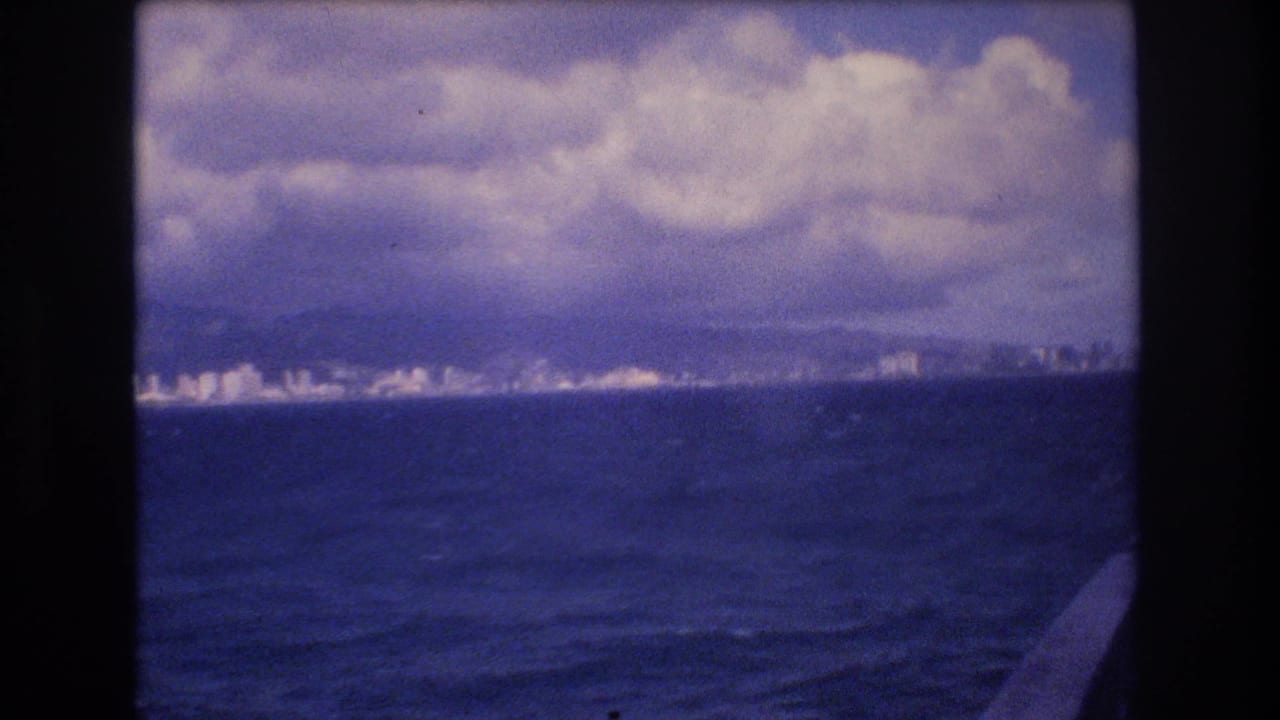 1977 8mm home-movie frame showing a distant Honolulu shoreline seen from a boat across dark blue water, with heavy clouds and a visible film border.