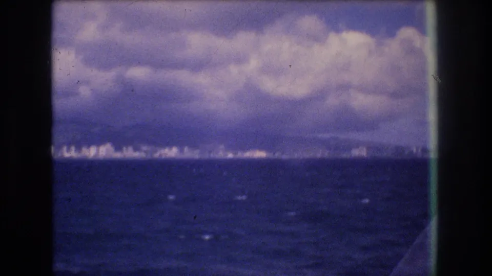 1977 Honolulu boat-view frame with open water in the foreground, a small distant coastal skyline, cloud cover, and dark 8mm frame edges.