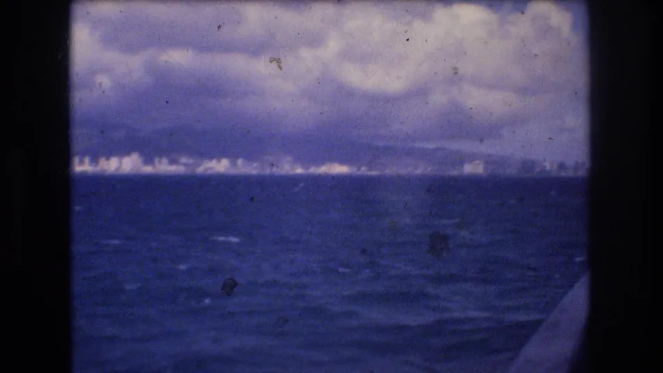 Soft 8mm frame of Honolulu's distant coastline across choppy blue water, with low clouds, haze, and the boat edge partly visible at right.