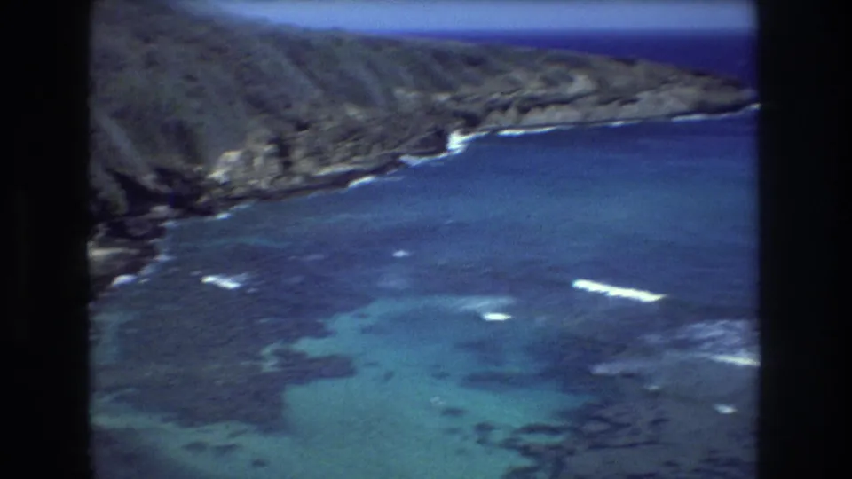 1977 Hawaii coastal frame showing rocky green cliffs above turquoise shallows, darker reef patches, white surf, and open blue water beyond.