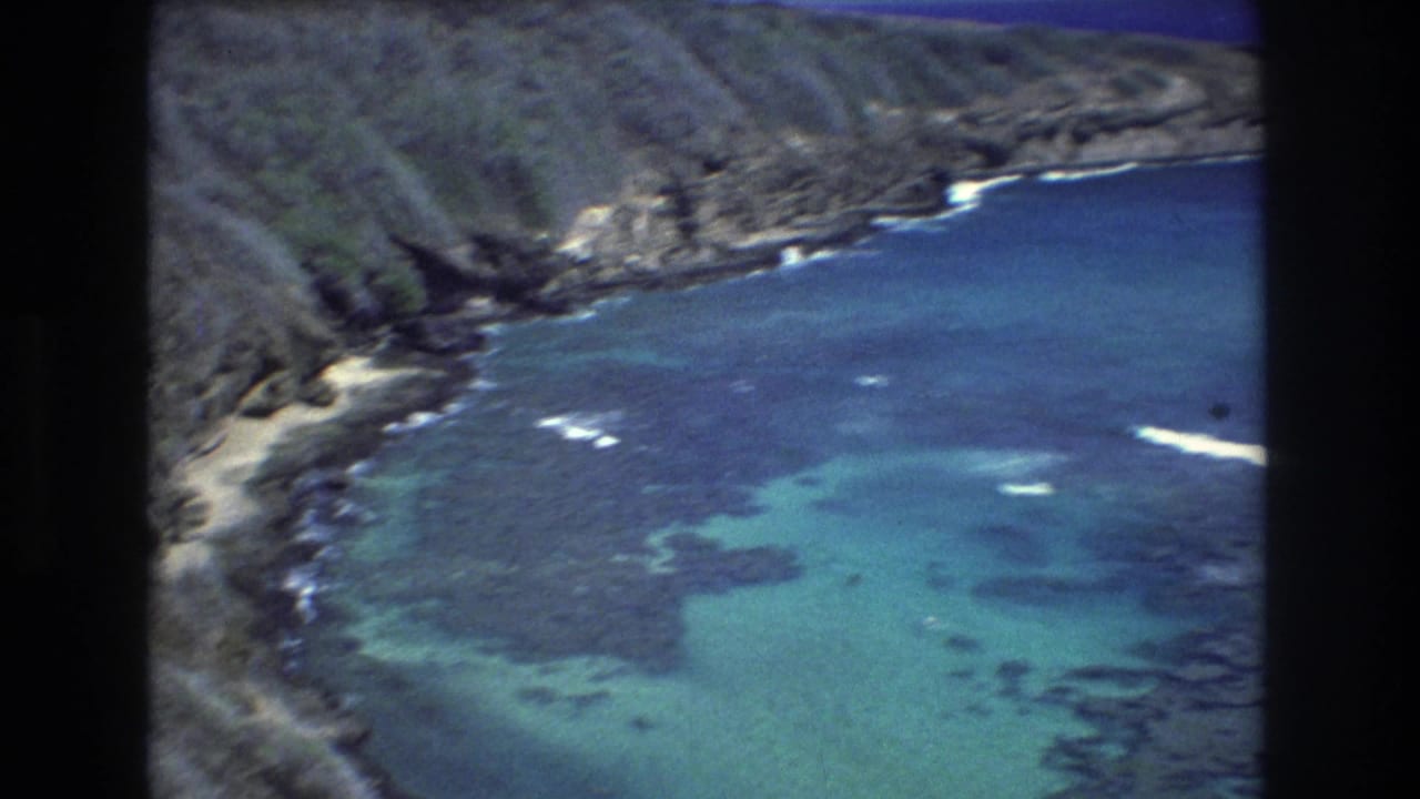 1977 8mm aerial view over a Honolulu, Hawaii shoreline, showing rugged coastal rock, shallow turquoise water, and dark submerged formations.