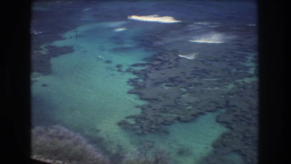 1977 8mm aerial frame over Honolulu coastal waters, with green-blue shallows, irregular dark rock pools, and small white surf breaks offshore.