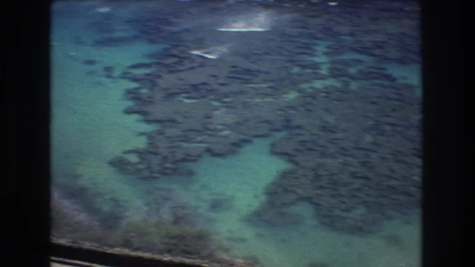 1977 8mm aerial view of Honolulu-area shallow sea water surrounding a broad, dark field of natural rock formations near the coast.