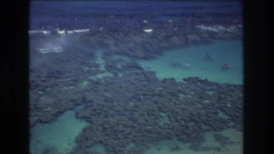 1977 8mm aerial frame showing a rocky Honolulu shoreline, a turquoise inlet or pool, and distant wave breaks along the coast.