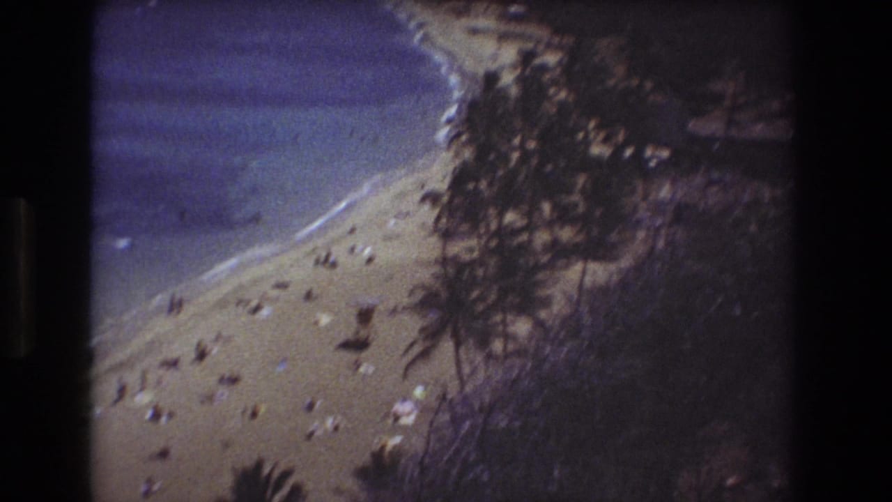 1977 8mm Honolulu-area beach view from above, showing scattered sunbathers on sand near blue water, with dark vegetation and hillside framing the shoreline.