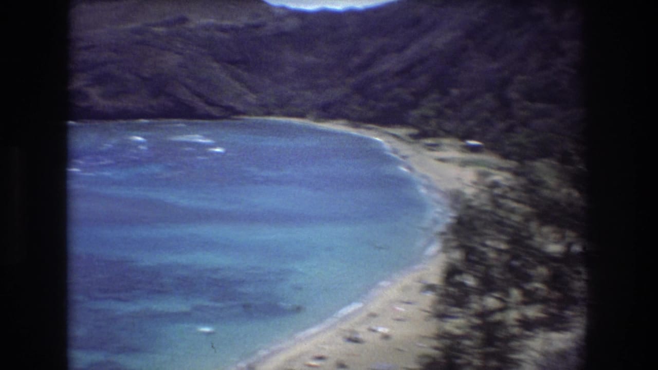Curved turquoise bay bordered by sand, vegetation, and dark green hills.