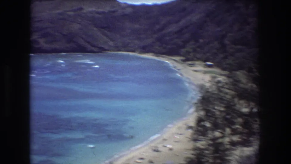 Wide 1977 8mm view of a Honolulu-area bay with turquoise water, a sandy curve of beach, coastal vegetation, and surrounding green hills.