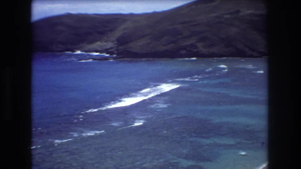 1977 8mm coastal view near Honolulu, showing blue water with white wave lines, a dark green hillside, and the edge of a sandy beach in the distance.
