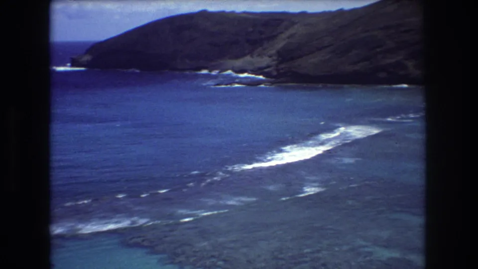 1977 8mm Honolulu-area coastline view with blue water, white breaking waves, rocky shore, and dark green hills under a pale sky.