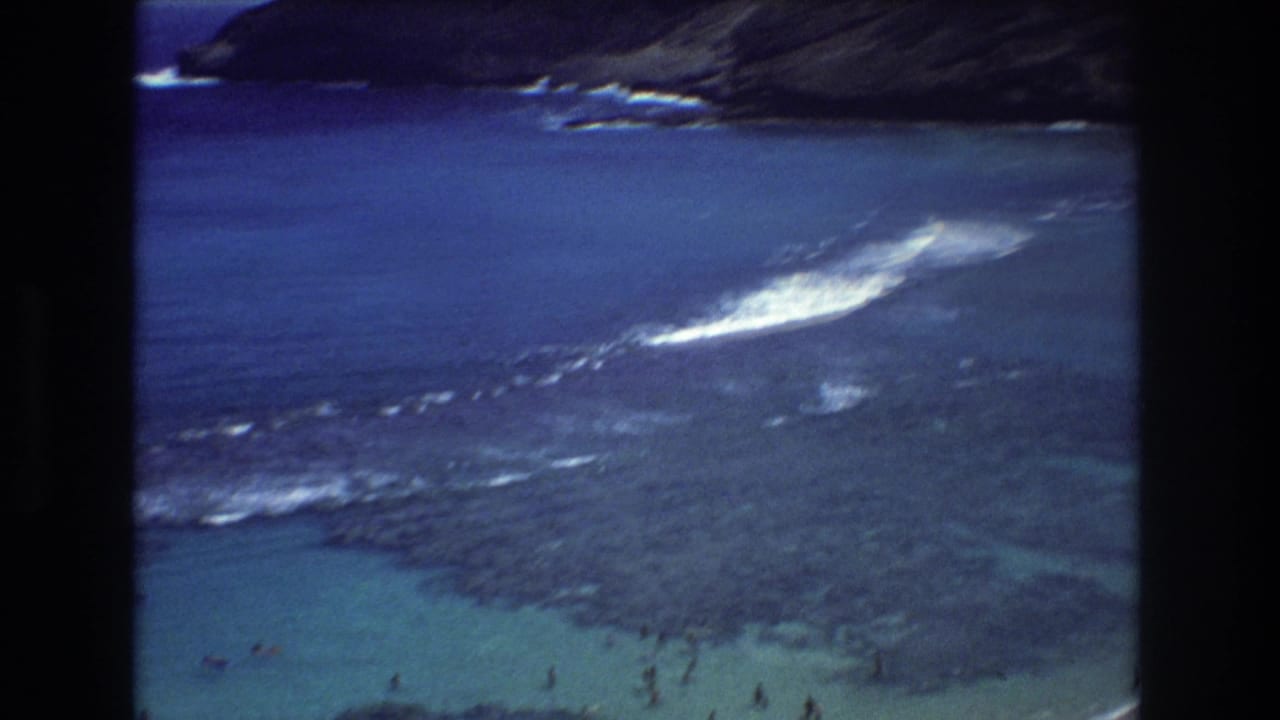 1977 Honolulu 8mm frame showing shallow turquoise water, dark reef rocks, white surf, distant bathers, and a coastal slope beyond the beach.