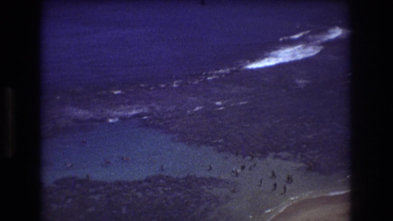 Rocky shoreline and shallow beach water with small groups of people near breaking waves.