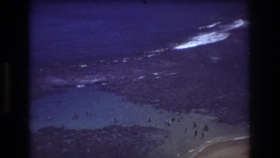 1977 Honolulu home-movie frame with distant people in the shallows, dark reef, white surf lines, and a sandy beach edge at lower right.
