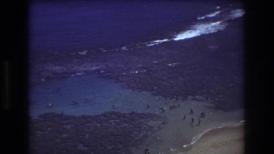 1977 Honolulu 8mm view of bathers near the shoreline, with reef rocks, pale sand, blue water, and white surf in the distance.