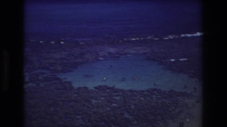 1977 Honolulu 8mm frame showing a wide rocky reef, blue ocean, turquoise tide-pool-like shallows, white wave lines, and distant people at the bottom edge.