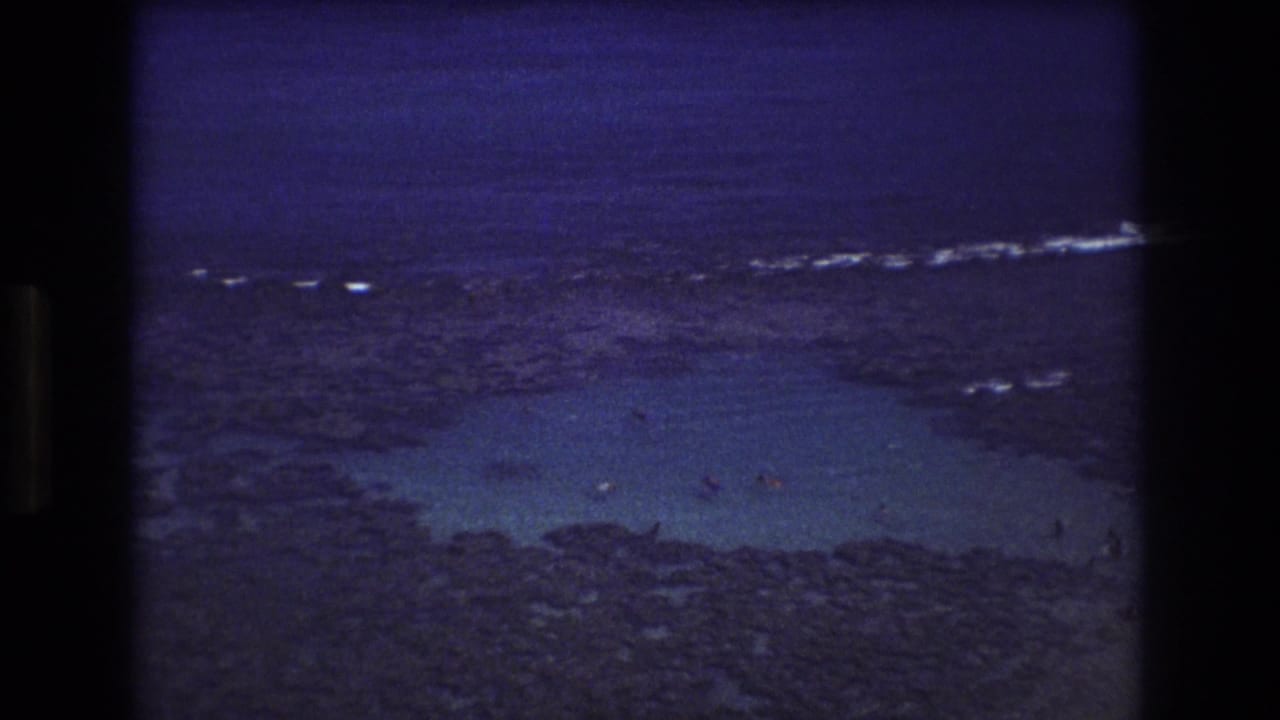 1977 Honolulu-area 8mm frame showing shallow turquoise shoals, dark rocky reef, distant surf, and small swimmers viewed from above.