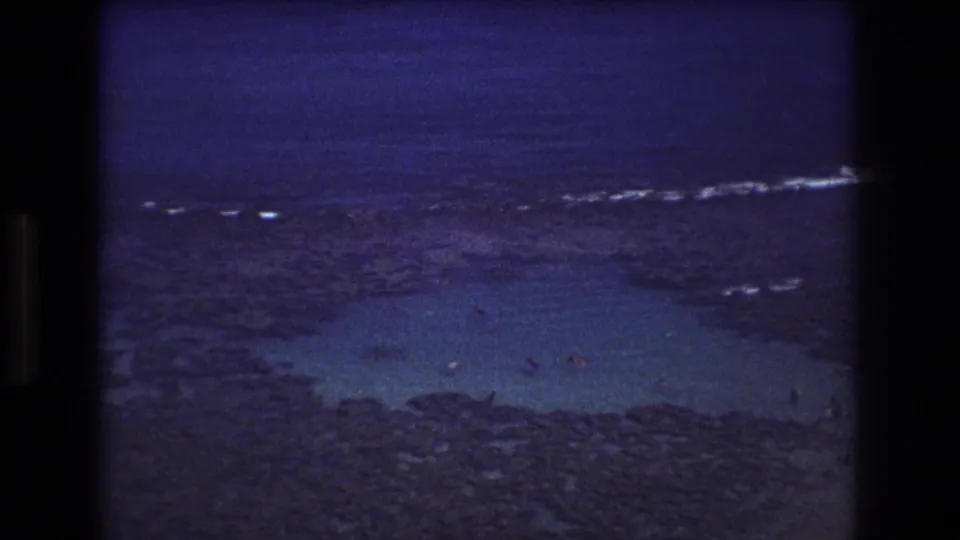 1977 Honolulu-area 8mm frame showing shallow turquoise shoals, dark rocky reef, distant surf, and small swimmers viewed from above.