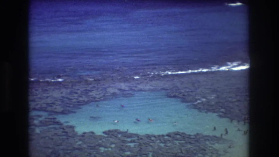 1977 Honolulu-area home-movie frame of a reef-protected turquoise pool with small swimmers, dark coral formations, and white surf beyond.