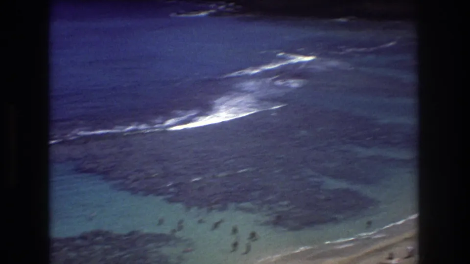1977 Honolulu-area 8mm view across shallow blue-green water, dark reef, small swimmers near the lower frame, and breaking surf offshore.