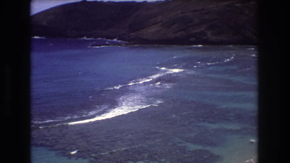 1977 Honolulu-area 8mm frame showing blue coastal water, dark reef, white surf breaks, and a large hillside in the background.