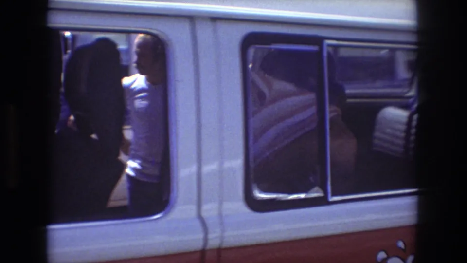 1977 Honolulu frame with a woman in a white T-shirt visible near the open side door of the red and white tour van, with other passengers partly obscured.