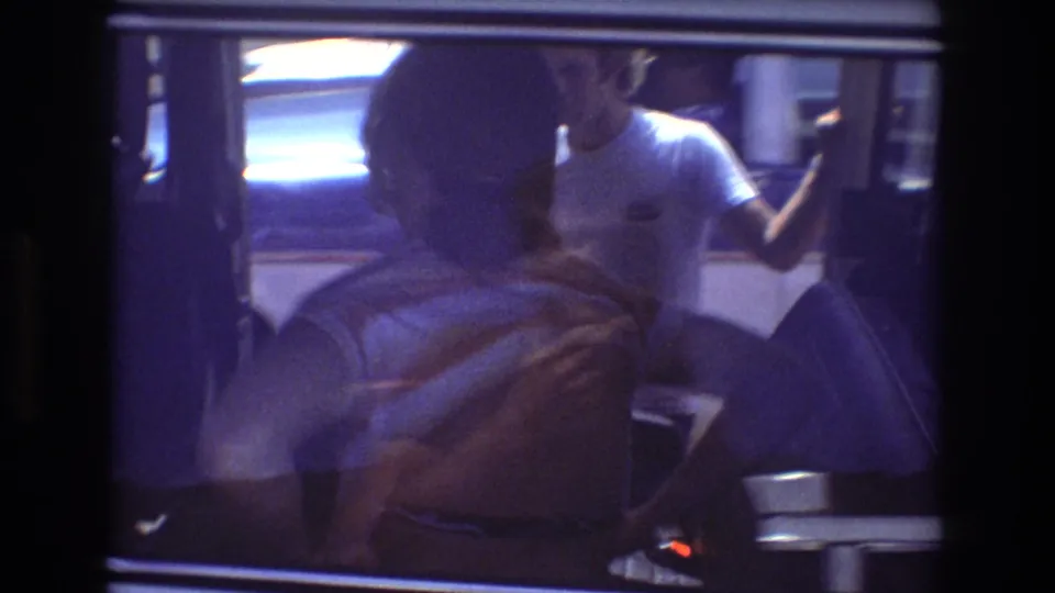 Soft 8mm frame from the Honolulu tour van sequence showing passengers through glass, including a woman in a white T-shirt near the open door and a closer obscured figure in the foreground.