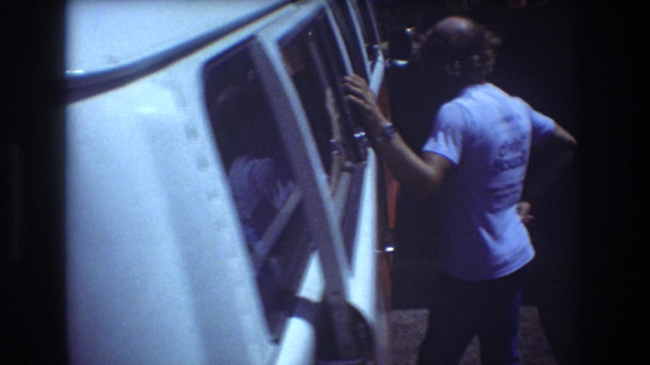 1977 Honolulu home-movie frame showing an adult in a light blue shirt standing close to the open side of a white or red-and-white van, with another person partly visible nearby.