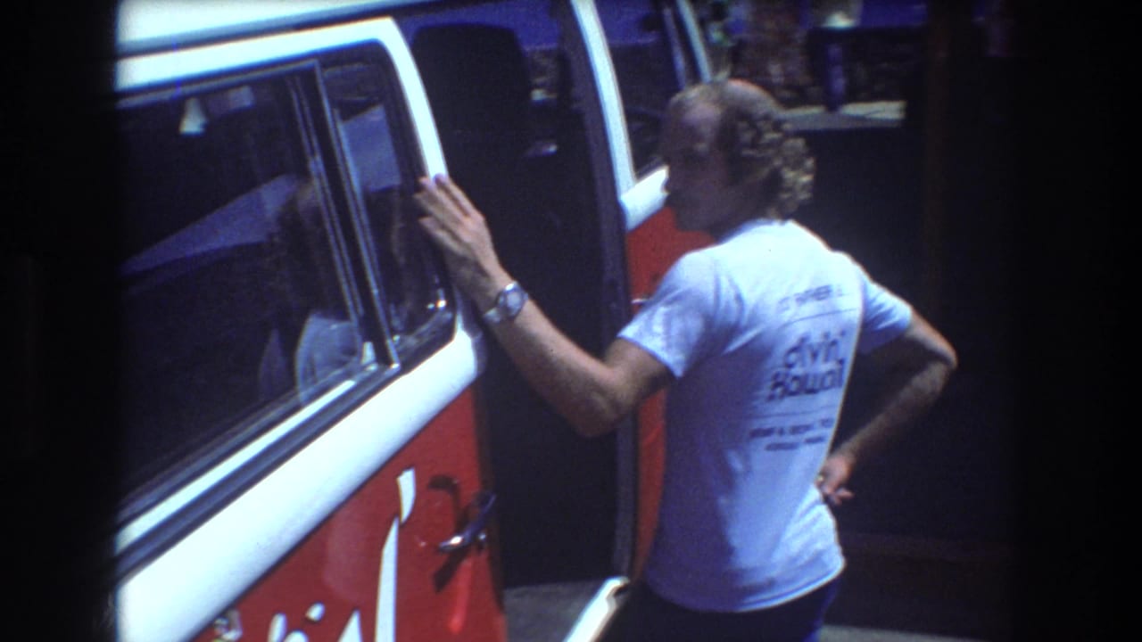 Adult in a light blue shirt leaning near a red-and-white van.