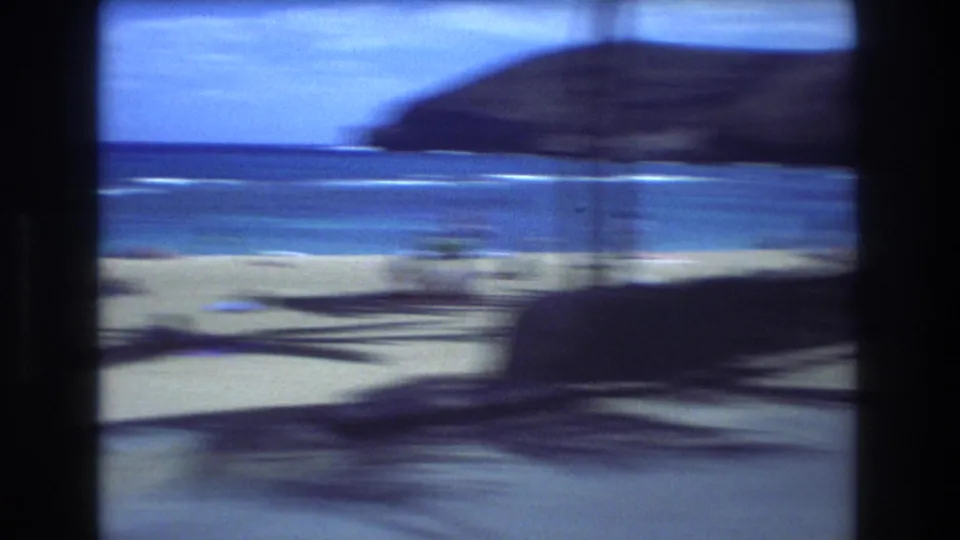 1977 Honolulu home-movie frame with strong motion blur across a beachside view, showing sand, ocean, and a dark foreground shape from the moving camera or vehicle.