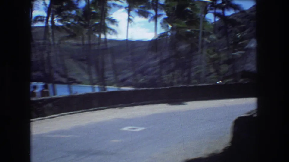 1977 Honolulu frame showing a road beside a beach area with palm trees, ocean water, and low hills in the distance, filmed from a moving handheld perspective.