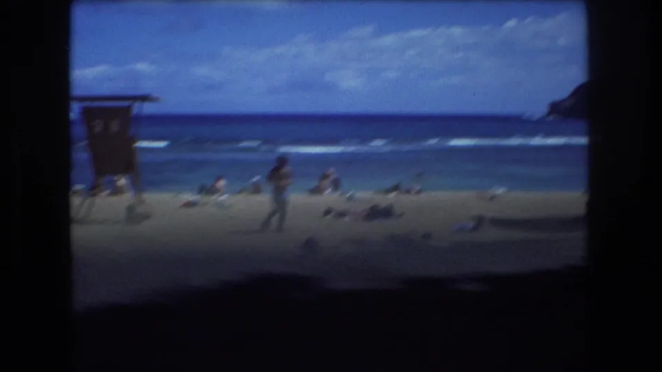 1977 Honolulu beach frame showing distant people on the sand, a small lifeguard-style structure, surf, blue ocean, and a hillside at the right edge.