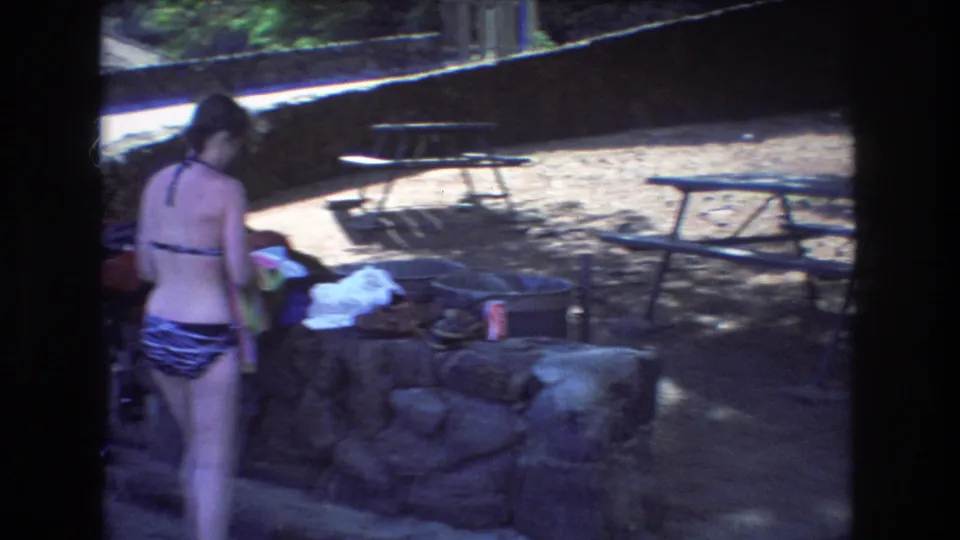 A woman in a patterned bikini stands at a stone picnic area in 1977 Honolulu footage, with towels, bags, metal picnic tables, and foliage nearby.