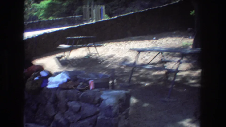 The frame shows a shaded Honolulu picnic area in 1977 with stonework, loose towels or clothing, a red beverage can, and metal tables under trees.