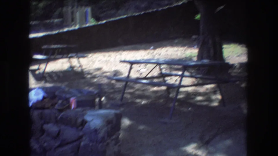 A closing frame from the Honolulu 1977 home movie shows metal picnic tables, stonework, scattered belongings, and heavy shade from nearby trees.